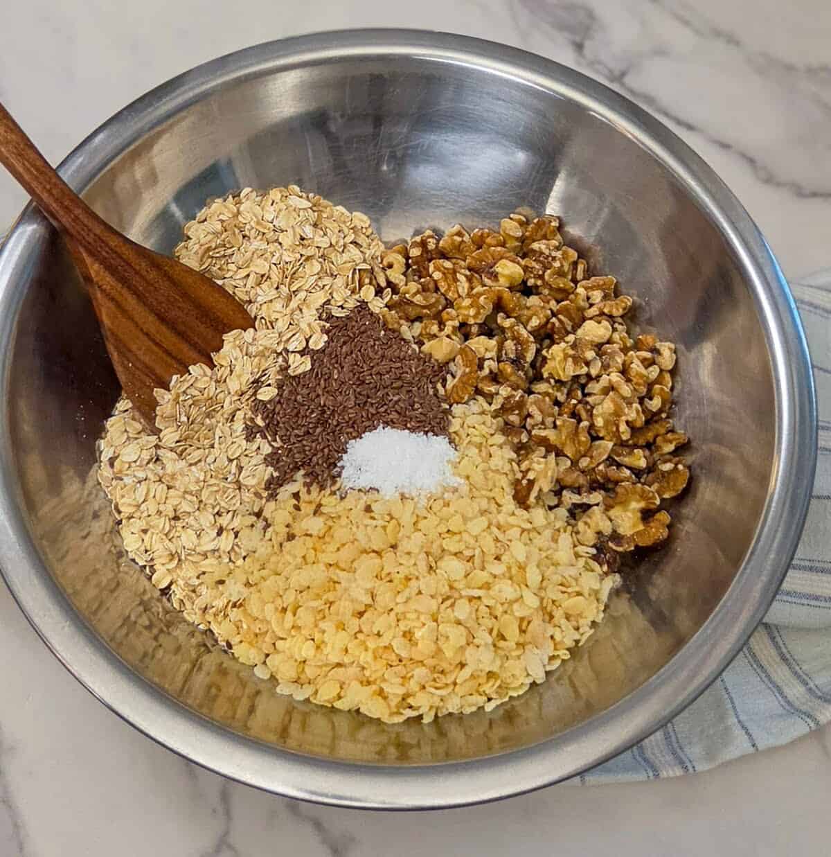 A large metal mixing bowl containing oats, rice cereal, walnuts, flax seeds, and salt for the banana walnut granola with a wooden spoon.