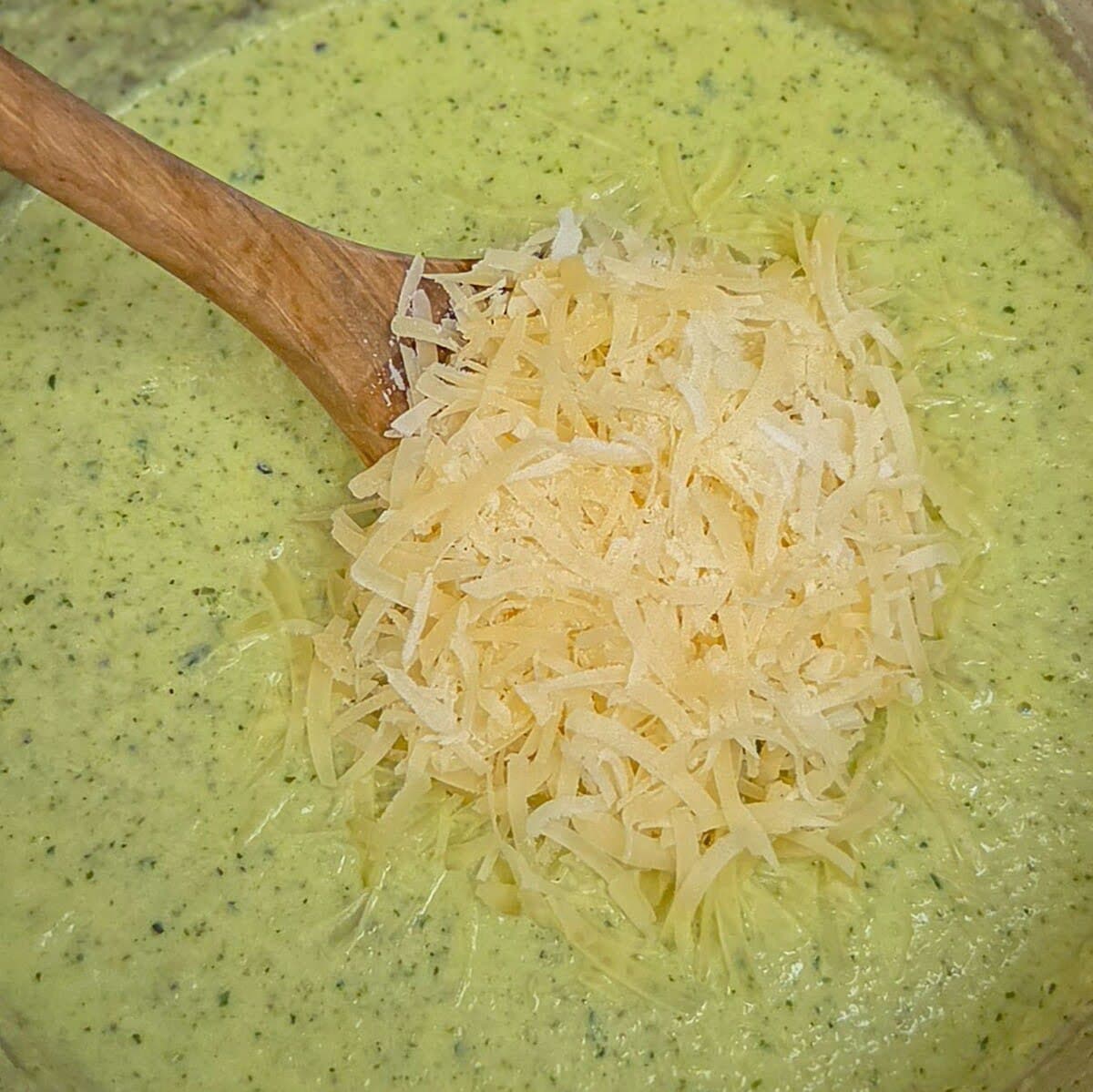 Adding shredded parmesan cheese to the creamy zucchini alfredo sauce in a saucepan with a wooden spoon.
