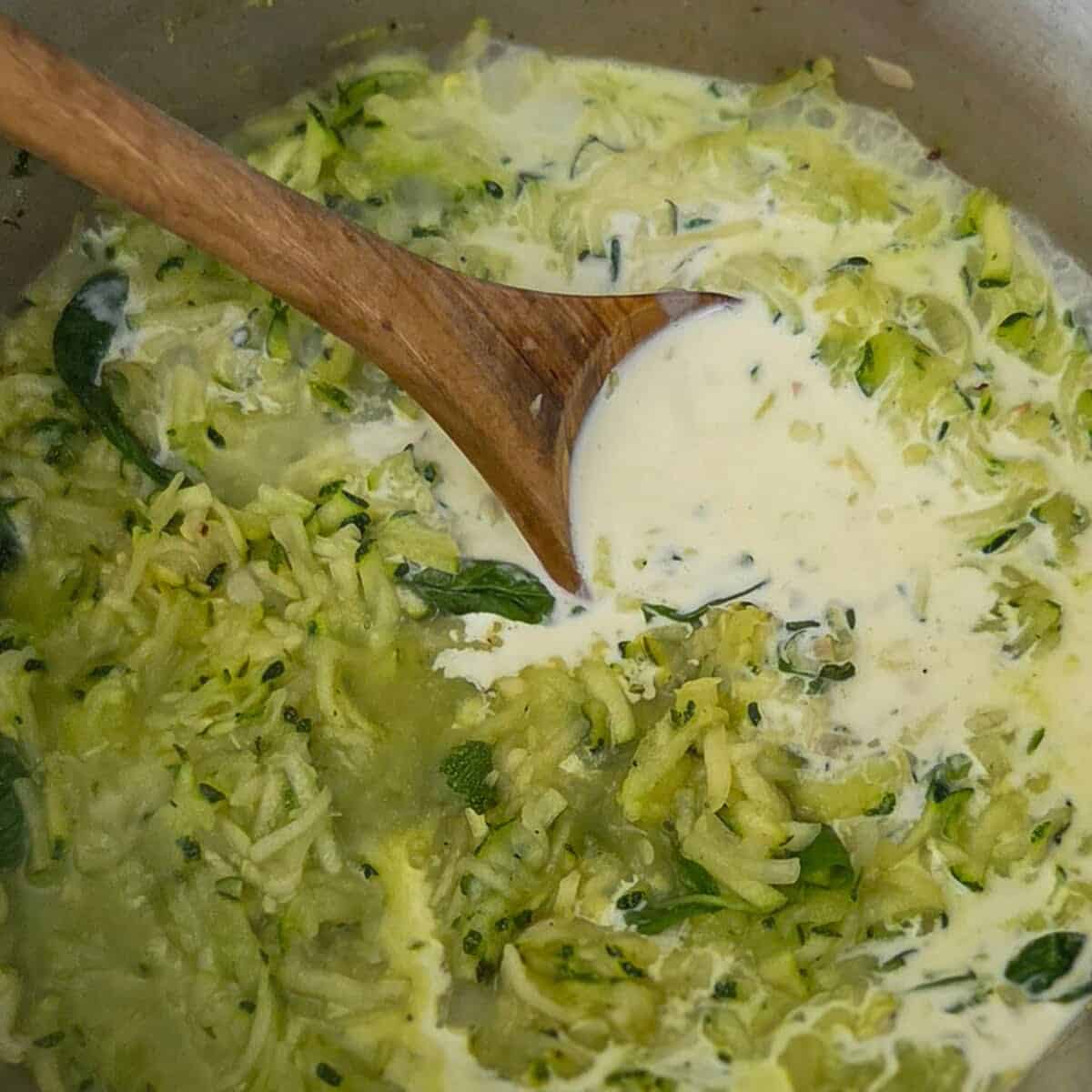 Adding cream to a saucepan containing shredded zucchini and basil leaves.