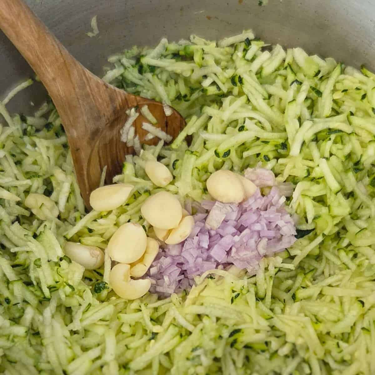 Adding garlic cloves and minced shallots to a saucepan with shredded zucchini using a wooden spoon.