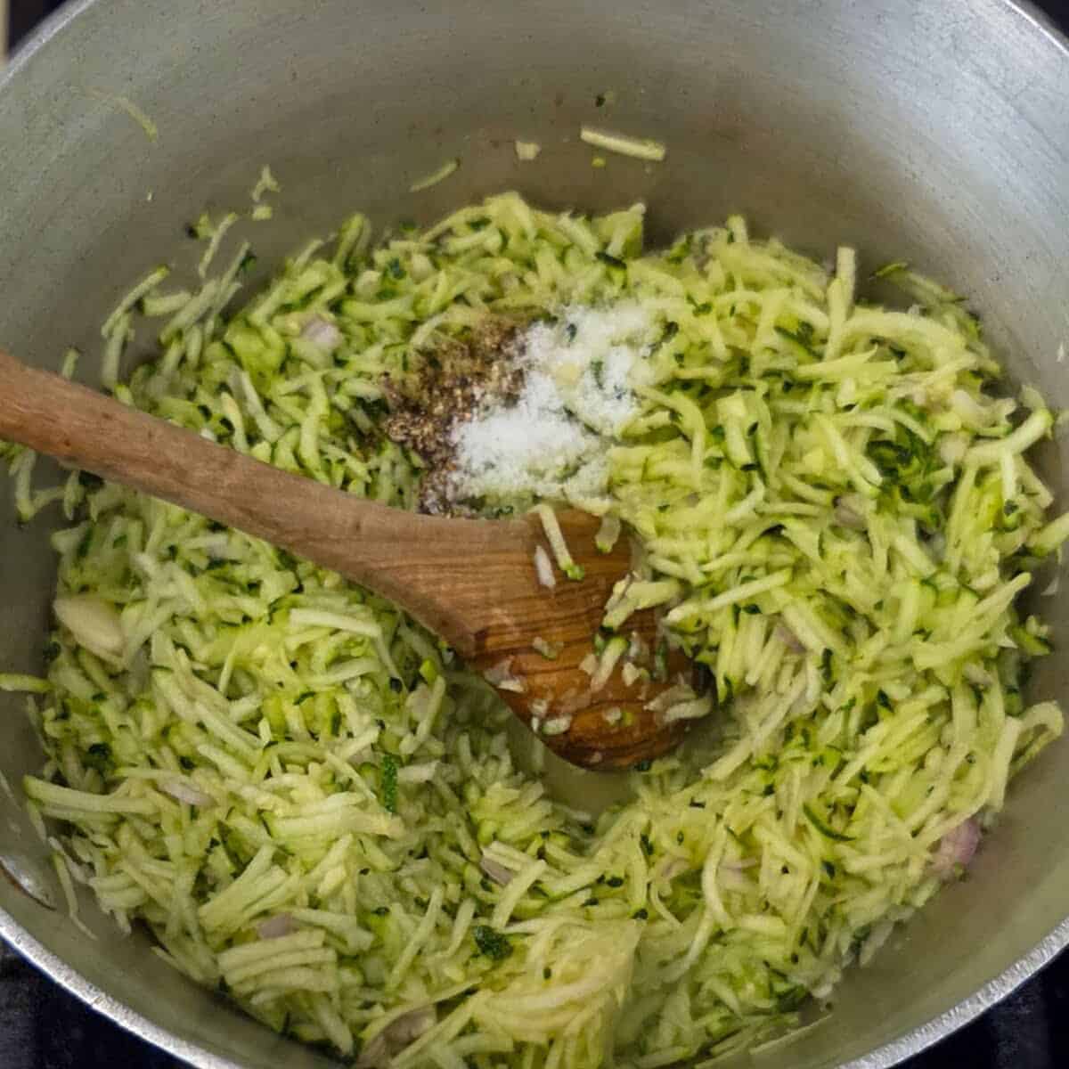Adding salt and pepper to a saucepan with shredded zucchini using a wooden spoon.