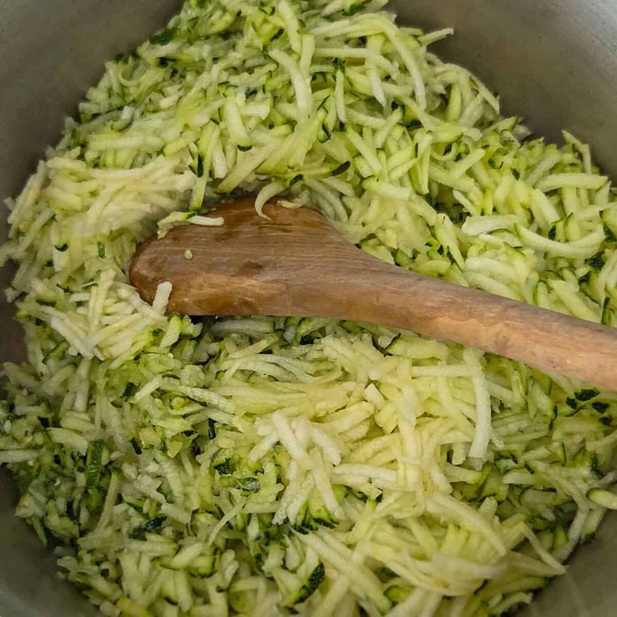 Adding the shredded zucchini to a saucepan using a wooden spoon.