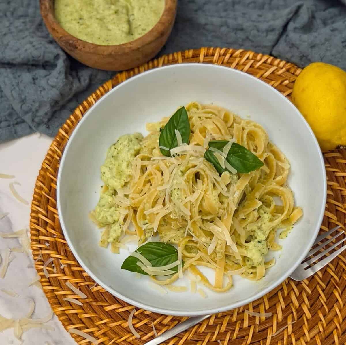 Creamy zucchini alfredo pasta on a plate with a lemon , a fork, and a bowl of sauce on the side.