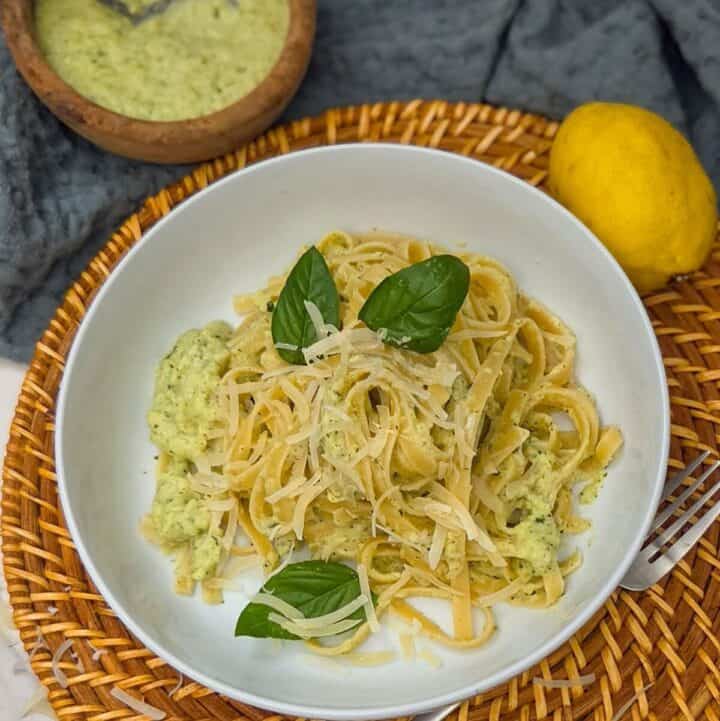 A plate of creamy zucchini alfredo pasta with a fork, and a bowl of sauce and a lemon on the side.
