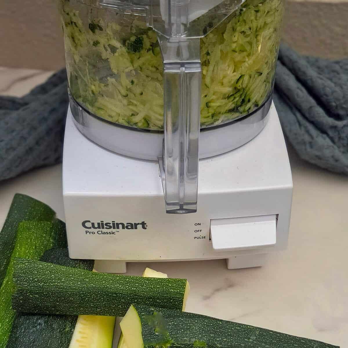Shredded zucchini in the bowl of a food processor with zucchini pieces laying next to it.