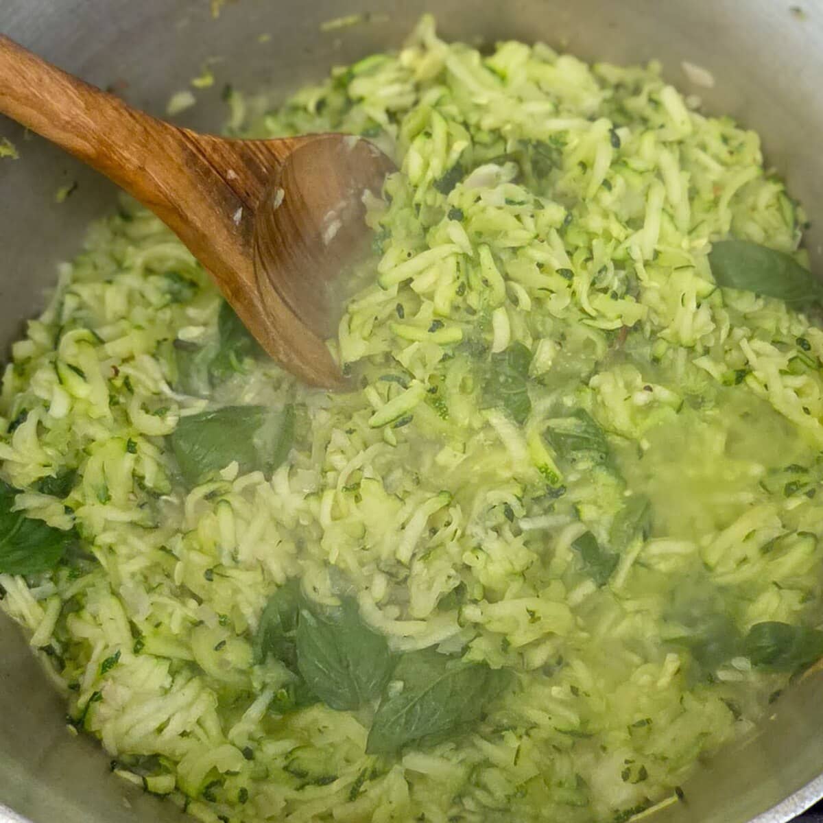 Stirring the shredded zucchini and basil leaves in a saucepan with a wooden spoon.